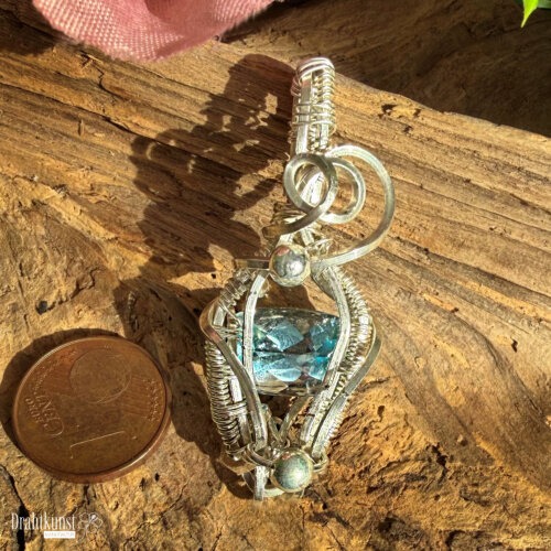 Handgefertigter Blautopas-Anhänger aus 935er Silber mit rechteckig facettiertem blauem Edelstein und geschwungener Drahtgestaltung, ca. 4,5 × 1,8 cm auf Holz